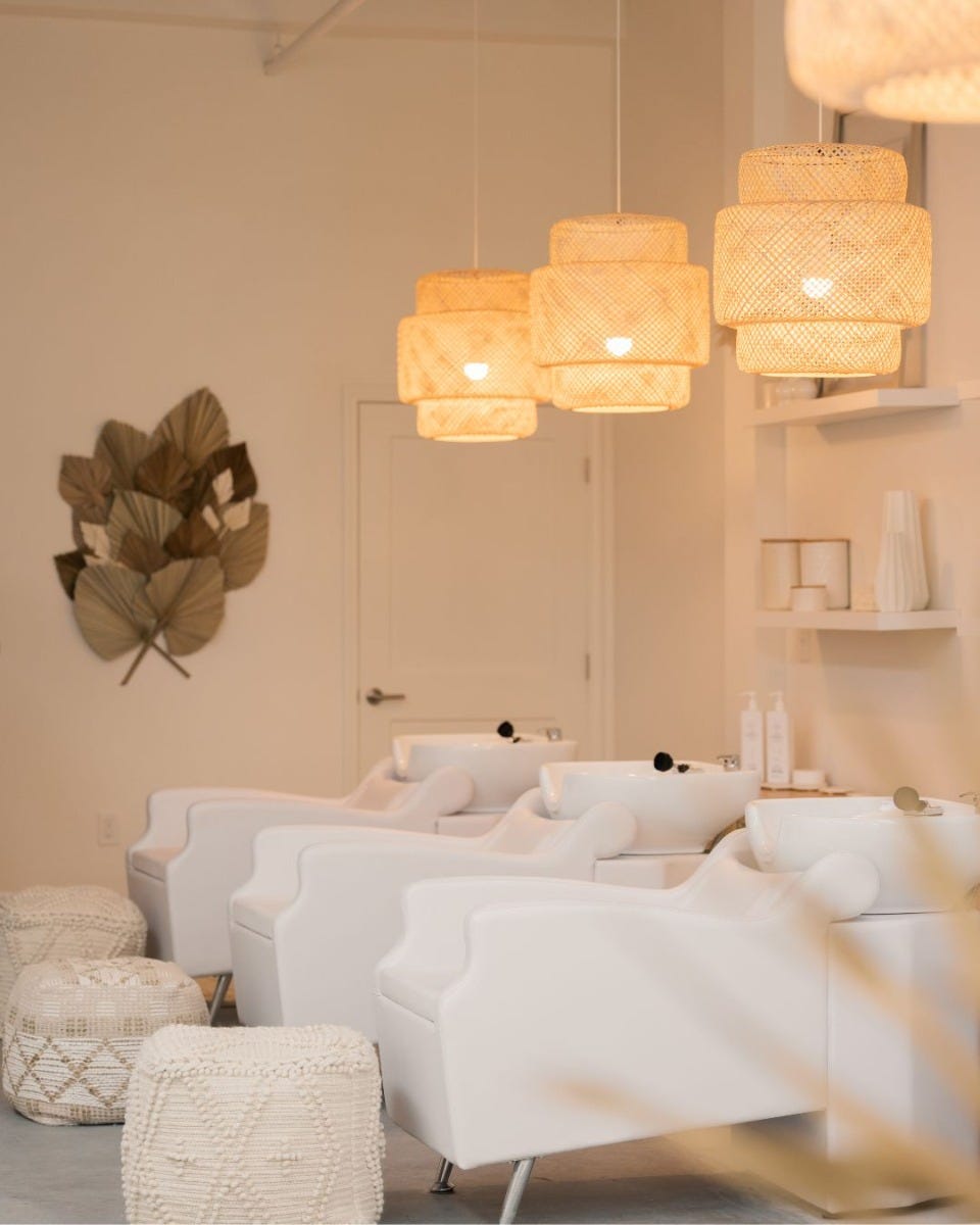 three all-white shampoo backwash stations with thatched pendant lighting and ottoman footrests