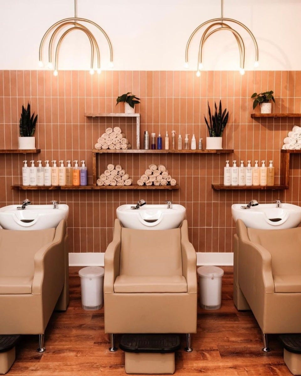 hair salon shampoo area with brown wall tiles, three camel shampoo backwash units and wall shelves with products and towels